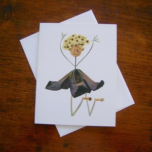 May include: A greeting card featuring a whimsical flower arrangement that resembles a person. The person is made of dried flowers and has a dark purple skirt, yellow flower hair, and green stems for arms and legs.