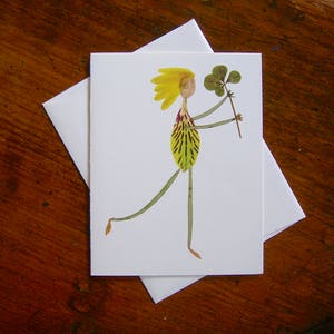 May include: A handmade greeting card featuring a whimsical figure made of pressed flowers. The figure has yellow hair, a green and yellow dress, and is holding a four-leaf clover.