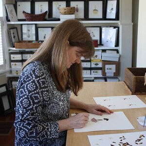 May include: A woman is working on a table with two pieces of paper. One piece of paper has a pressed flower arrangement and the other has pressed leaves and flowers. The table is made of wood and has a light brown finish.
