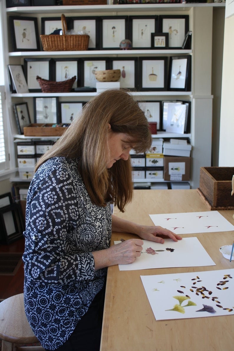 May include: A woman is working on a table with two pieces of paper. One piece of paper has a pressed flower arrangement and the other has pressed leaves and flowers. The table is made of wood and has a light brown finish.