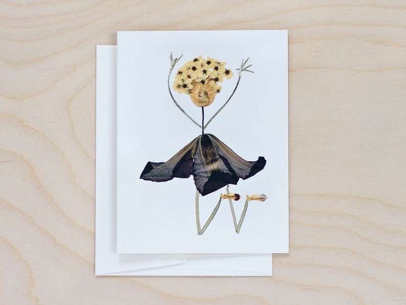 May include: A greeting card featuring a whimsical figure made of pressed flowers. The figure has a yellow flower crown, a dark purple dress, and outstretched arms.