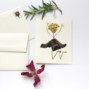 May include: A handmade greeting card featuring a pressed flower arrangement in the shape of a person. The person has a yellow flower head, a brown stem body, and a dark purple flower skirt. The card is blank inside for writing a message.