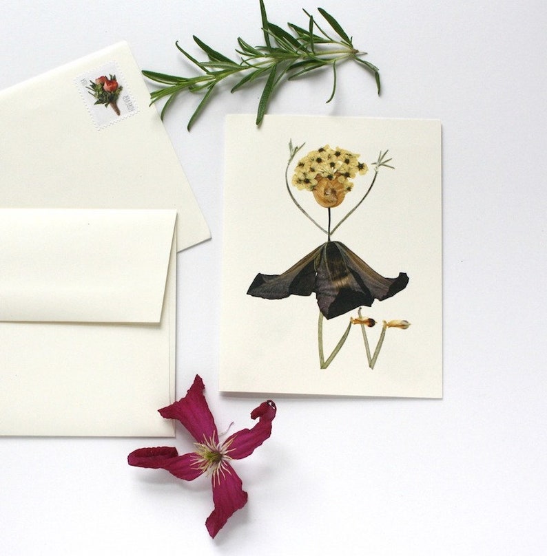 May include: A handmade greeting card featuring a pressed flower arrangement in the shape of a person. The person has a yellow flower head, a brown stem body, and a dark purple flower skirt. The card is blank inside for writing a message.