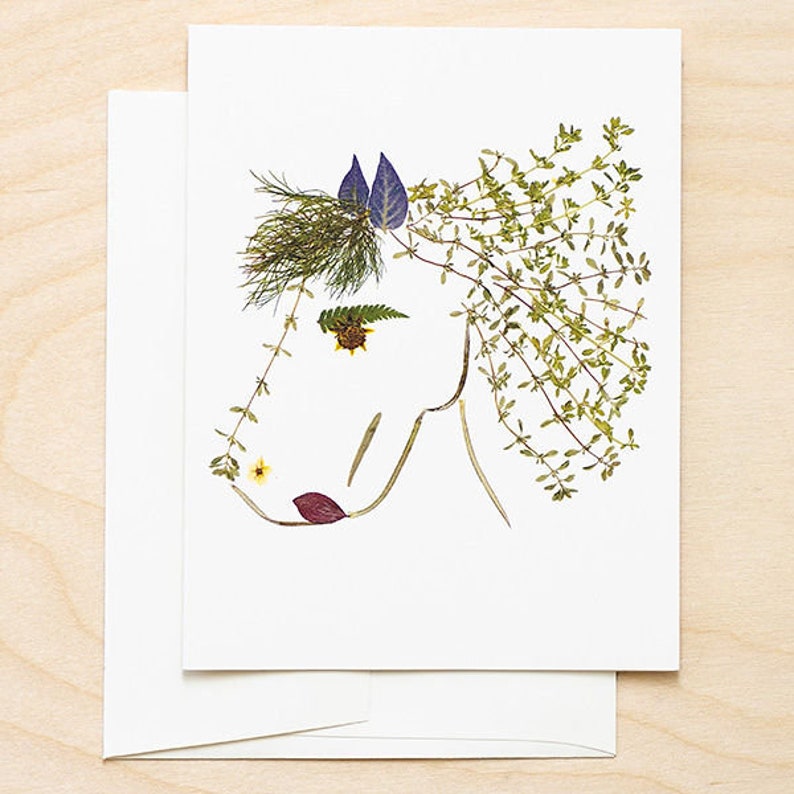 May include: A white card with a horse's head made from pressed flowers and leaves. The horse's mane is made from green leaves, the ears are purple, and the nose is a yellow flower.