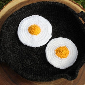 May include: A black crocheted skillet with a handle, holding two crocheted fried eggs. The eggs have white 'whites' and yellow 'yolks'. The skillet rests on a wooden surface, with green grass visible in the background.
