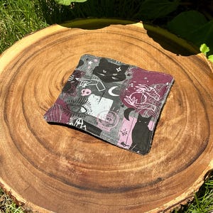 May include: Square fabric coaster with a black cat, moon, eye, and other mystical symbols on a gray and purple background. The coaster is placed on a wooden surface.