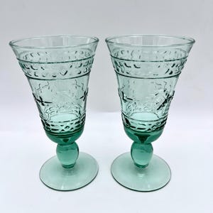 May include: Two vintage green-tinted glass goblets. Each goblet features a wide, textured rim and a detailed pattern on the body. The stem is a rounded ball shape, and the base is a flat, circular design. The goblets are displayed against a white background.