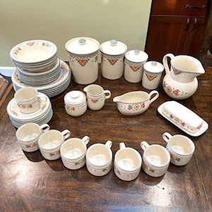 May include: A complete cream-colored dinnerware set with a Southwestern design. The set includes plates, bowls, cups, saucers, serving dishes, a creamer, sugar bowl, and a pitcher. The set has a geometric pattern in shades of red, orange, and blue.
