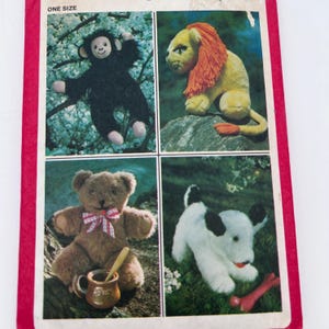 May include: A sewing pattern envelope featuring four stuffed animals: a black monkey, a yellow lion with an orange mane, a brown teddy bear wearing a red and white plaid bow tie with a honey pot, and a white and black puppy with a red bone toy.