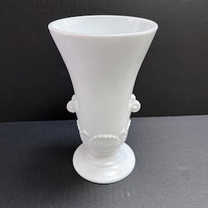 May include: A white milk glass vase featuring a flared rim, a pedestal base, and decorative molded details including side scrolls and a shell-like pattern around the lower body. The vessel has a smooth, glossy finish and a wide, open top.