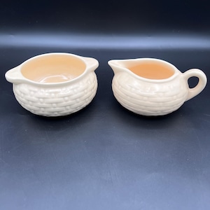 May include: Two-piece cream-colored ceramic serving set. The set includes a small bowl with handles and a creamer pitcher with a handle and spout. Both pieces have a woven basket-like texture on the exterior and a peach-colored interior.