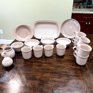 May include: A collection of light pink ceramic dinnerware, including plates, bowls, mugs, and serving dishes. The set includes a large oval platter, several round plates, and various sized bowls. A salt shaker and a creamer are also included.
