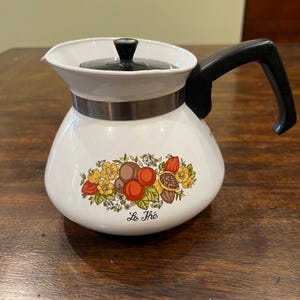 May include: White enamel teapot with a black handle and lid. The teapot features a decorative floral and fruit design with the words "Le Thé" printed on the front. The teapot has a silver band around the middle.