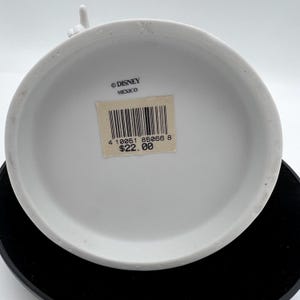 May include: A white ceramic Disney figurine base with a sticker that reads "&copy; DISNEY MEXICO". The sticker also has a barcode and the number "85066". The figurine is on a black surface.