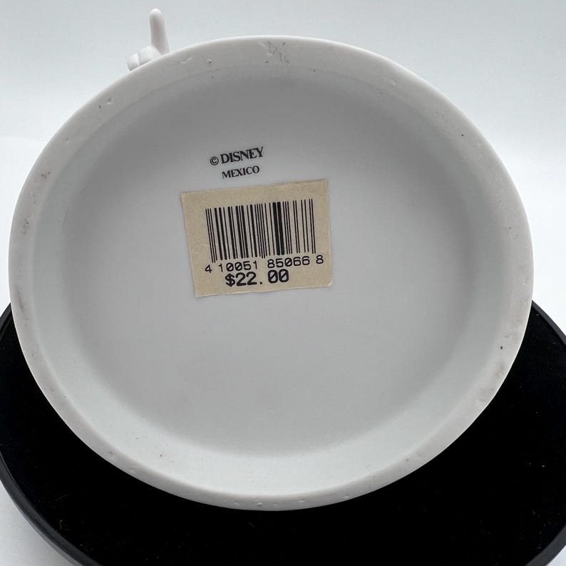 May include: A white ceramic Disney figurine base with a sticker that reads "&copy; DISNEY MEXICO". The sticker also has a barcode and the number "85066". The figurine is on a black surface.