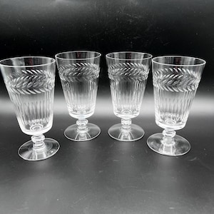 May include: A set of four clear glass goblets featuring a horizontal etched laurel leaf pattern around the upper rim and vertical fluted detailing along the lower bowl. Each glass stands on a short, faceted stem with a circular base.