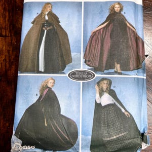 May include: A sewing pattern envelope by Teresa Nordstrom featuring four variations of capes and cloaks. The images display long, hooded, and shoulder-draped outerwear in brown, black, and deep red fabrics, worn over dresses and skirts.