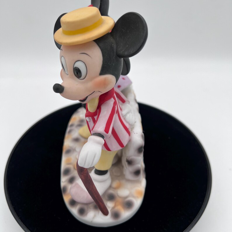 May include: A vintage Mickey Mouse figurine. Mickey is wearing a red and white striped jacket, yellow trousers, a yellow hat, and white gloves. He is holding a cane and standing on a decorative base with a spotted design.