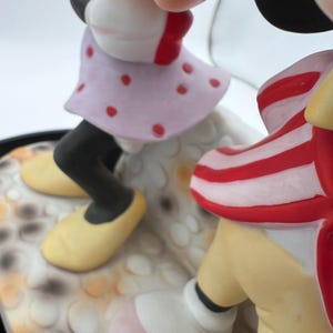 May include: A ceramic figurine of Minnie and Mickey Mouse. Minnie is in a light purple dress with red polka dots, black leggings, and yellow shoes. Mickey wears red and white striped shorts and yellow shoes. The figurine has a white and tan base.