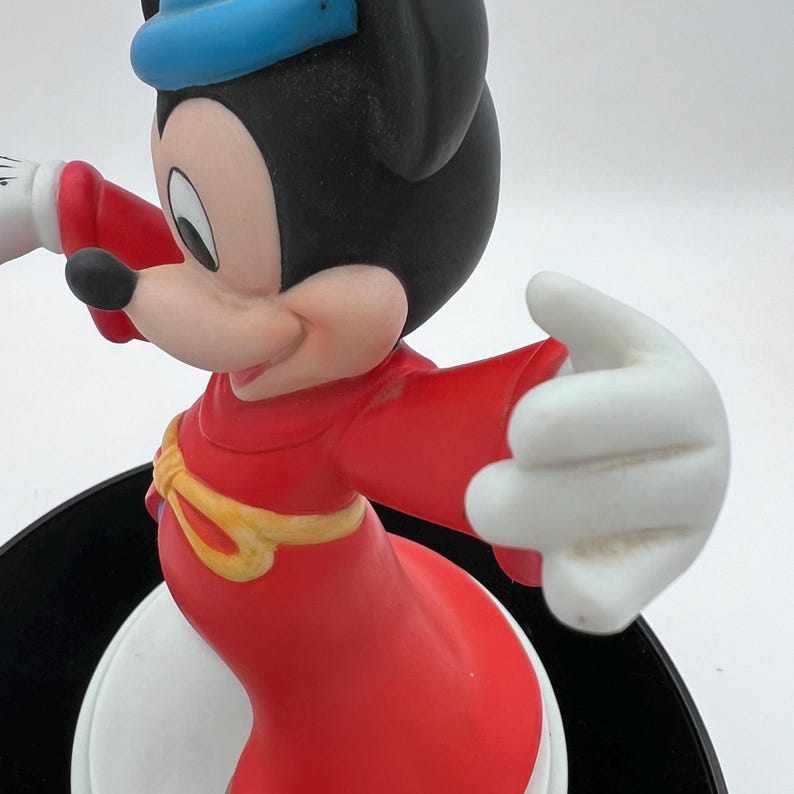 May include: A figurine of Mickey Mouse in a red robe and blue hat. The figure has white gloves and a yellow belt. The base is black and white. The figurine is a collectible item.
