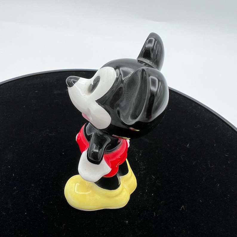 Peut inclure: Figurine en c&eacute;ramique de Mickey Mouse. Le personnage a un corps noir, un visage blanc et un short rouge. Il porte des chaussures jaunes et est pr&eacute;sent&eacute; dans une pose classique, mettant en valeur le design embl&eacute;matique.