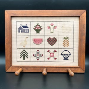 May include: A framed cross-stitch sampler with a wooden frame and three hooks on the bottom. The sampler features a variety of traditional designs, including a house, a goose, a watermelon, a heart, a pineapple, a tree, a quilt block, a flower, and a basket.