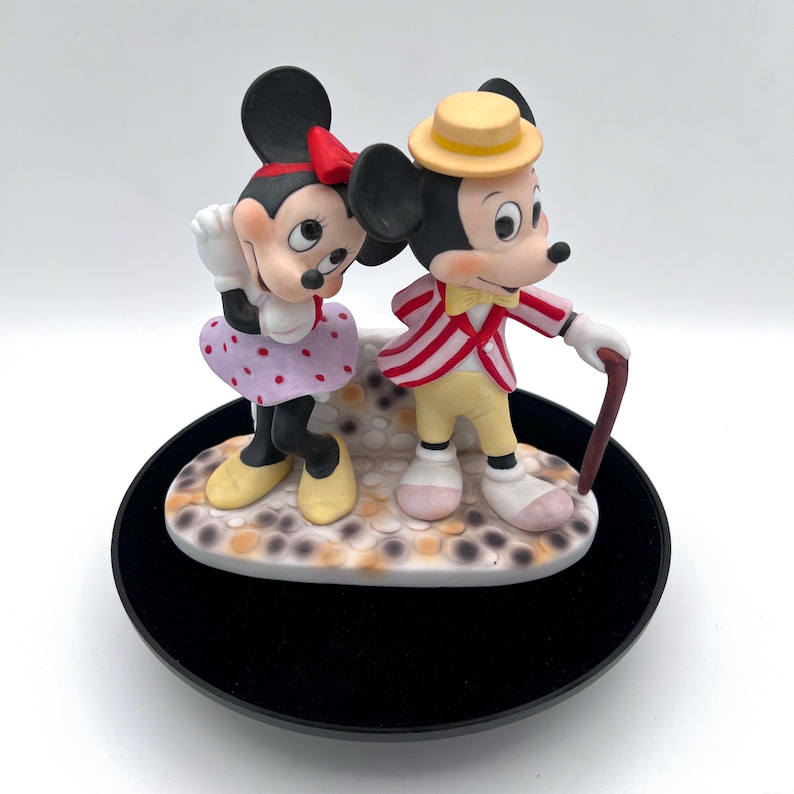 May include: A ceramic figurine of Mickey and Minnie Mouse. Minnie wears a red bow, polka dot skirt, and yellow shoes. Mickey wears a yellow hat, red and white striped jacket, and carries a cane. The figurine sits on a decorative base.