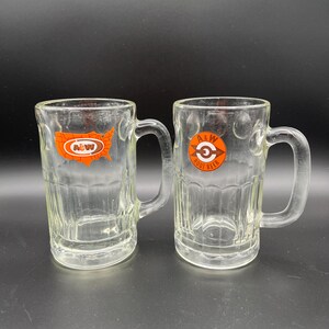 May include: Two clear glass mugs with handles. The mug on the left has a red and orange A&W logo with a map of the United States. The mug on the right has a red and orange A&W Root Beer logo.