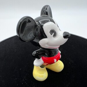 Peut inclure: Figurine en c&eacute;ramique de Mickey Mouse, avec une t&ecirc;te et des oreilles noires brillantes, un visage blanc et un short rouge. La figurine a des chaussures jaunes et est en position de marche. La figurine est sur une surface noire.