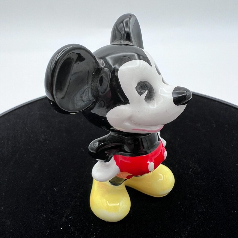 Peut inclure: Figurine en c&eacute;ramique de Mickey Mouse, avec une t&ecirc;te et des oreilles noires brillantes, un visage blanc et un short rouge. La figurine a des chaussures jaunes et est en position de marche. La figurine est sur une surface noire.