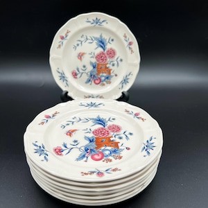 May include: A stack of white ceramic plates with scalloped edges. Each plate features a floral design with pink flowers, blue leaves, and orange accents. The plates are stacked on a black surface.