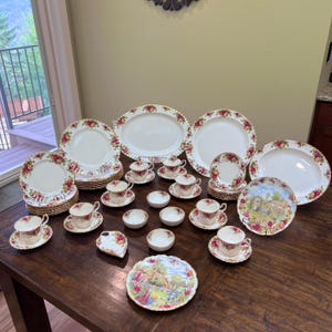 May include: A collection of vintage china with a floral rose pattern. The set includes plates, saucers, teacups, serving platters, and small bowls. The china has a white base with a gold trim.