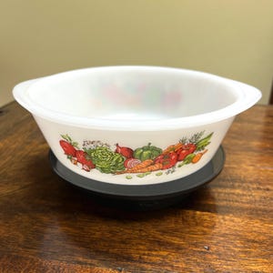 May include: White Pyrex bowl with a colorful vegetable design. The bowl features a variety of vegetables including tomatoes, carrots, lettuce, and onions. The bowl is sitting on a black base and has two handles.