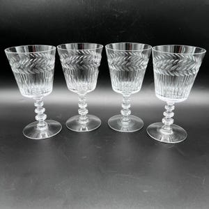 May include: A set of four clear glass goblets featuring a horizontal etched laurel leaf pattern around the upper rim and vertical fluted detailing on the lower bowl. Each glass has a multi-tiered, ball-shaped stem and a circular base.