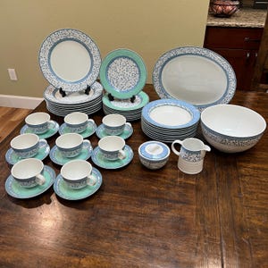 May include: A complete dinnerware set featuring plates, bowls, cups, saucers, and serving dishes. The set has a white base with a blue floral pattern and a teal green border. Includes a large oval platter, a sugar bowl, and a creamer.