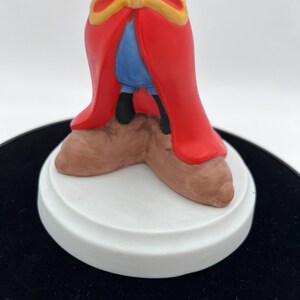 May include: A ceramic figurine featuring a character in a red robe with gold trim, blue pants, and brown shoes. The figurine is mounted on a white circular base. The figure's pose and attire suggest a fantasy or fairytale theme.