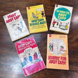 May include: Five vintage Andy Capp comic books are displayed on a wooden surface. The books feature colorful covers with illustrations and text, including titles like "Meet Andy Capp" and "Andy Capp Strikes Back."