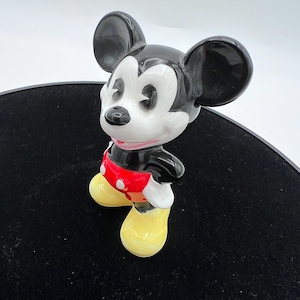 Peut inclure: Figurine en c&eacute;ramique de Mickey Mouse. La figurine a des oreilles noires, un corps noir et un visage blanc. Mickey porte un short rouge &agrave; boutons blancs et des chaussures jaunes. La figurine est pos&eacute;e avec les mains sur les hanches.