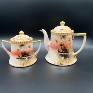 May include: A vintage tea set with a delicate floral design. The teapot and sugar bowl are both white with gold accents and feature a hand-painted scene of a lake with swans and a small house in the background.