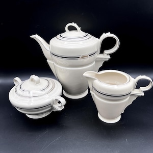 May include: A three-piece porcelain tea set in cream with silver trim accents. The set includes a lidded teapot, a lidded sugar bowl with two side handles, and a creamer pitcher. Each piece features a smooth, rounded body and a matching silver band.