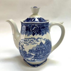 May include: A white and blue ceramic teapot with a lid. The teapot features a blue landscape scene with a windmill and floral patterns. The handle and spout are white. The lid has a floral knob.