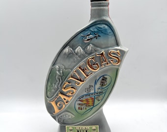 May include: A gray ceramic bottle with a Las Vegas theme. The bottle features a gold-colored "LAS VEGAS" inscription, illustrations of a helicopter, mountains, and other Vegas icons. The bottle has a rectangular label at the bottom.