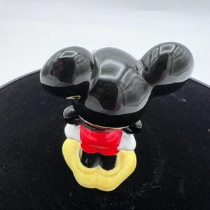 Peut inclure: Figurine en c&eacute;ramique de Mickey Mouse, vue de dos. La figurine a de grandes oreilles noires brillantes, un short rouge, des gants blancs et des chaussures jaunes. La figurine est sur une surface noire.