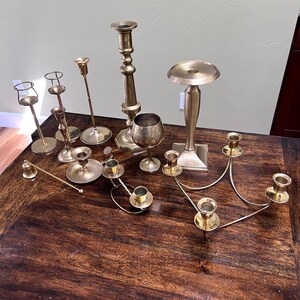 May include: A collection of various brass candle holders and a brass goblet. The assortment includes tall tapered candlesticks, a multi-armed candelabra, a short candle snuffer, and a pedestal-style candle stand, all featuring a metallic gold finish.