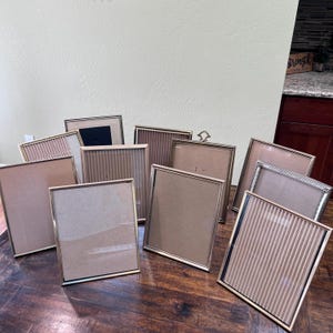 May include: A collection of ten rectangular picture frames with thin metallic gold-toned borders. The frames feature various internal designs, including smooth glass surfaces and textured, vertically ribbed inserts in neutral beige and tan tones.