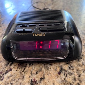 May include: Black Timex digital alarm clock displaying 1:17 in red numerals. Features AM/FM radio frequency indicators and buttons for time, alarm, and snooze functions. The clock is on a speckled countertop.