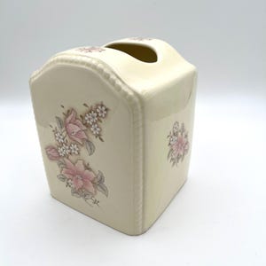 May include: Cream-colored ceramic tissue box with a floral design. The box has a rectangular shape with a curved top and a decorative beaded edge. Pink and white flowers with gray leaves adorn the sides, adding a touch of elegance.