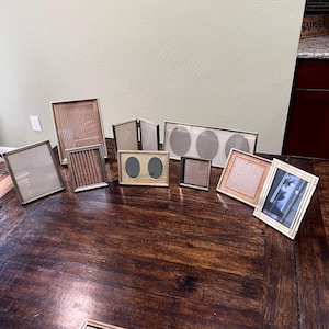May include: A collection of various metal and wood picture frames in different shapes and sizes, including rectangular, square, and oval openings. The frames feature metallic, silver, and light wood finishes, arranged on a dark wooden surface.