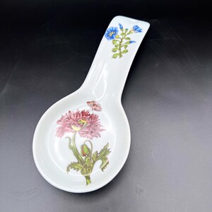 May include: White ceramic spoon rest with floral and butterfly designs. The spoon rest features a large red and pink flower with green leaves and a small butterfly. Blue flowers adorn the handle.
