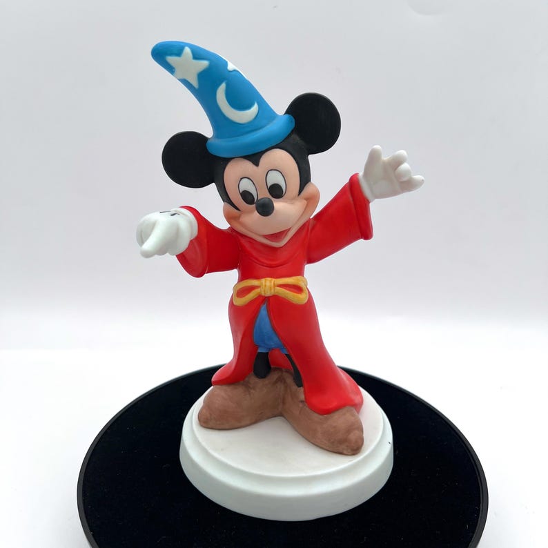 May include: A figurine of Mickey Mouse dressed as a sorcerer, wearing a blue hat with a crescent moon and star, a red robe, and brown shoes. The figure is standing on a white base with a black circular stand.